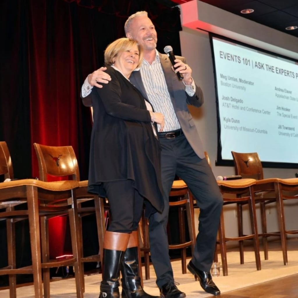 Two colleagues embracing on stage while speaking in to the microphone.