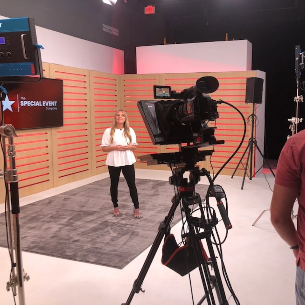 A women in business casual standing on a set speaking in to a camera with The Special Event Company logo on a tv in the background.