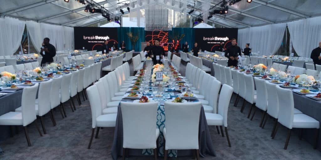 Dining room with multiple long tables with white cloth chairs and blue table settings.