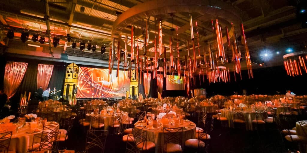 Ballroom set up for an elegant dinner with multiple round tables and chairs with place settings. Unique chandeliers hang from the ceiling.